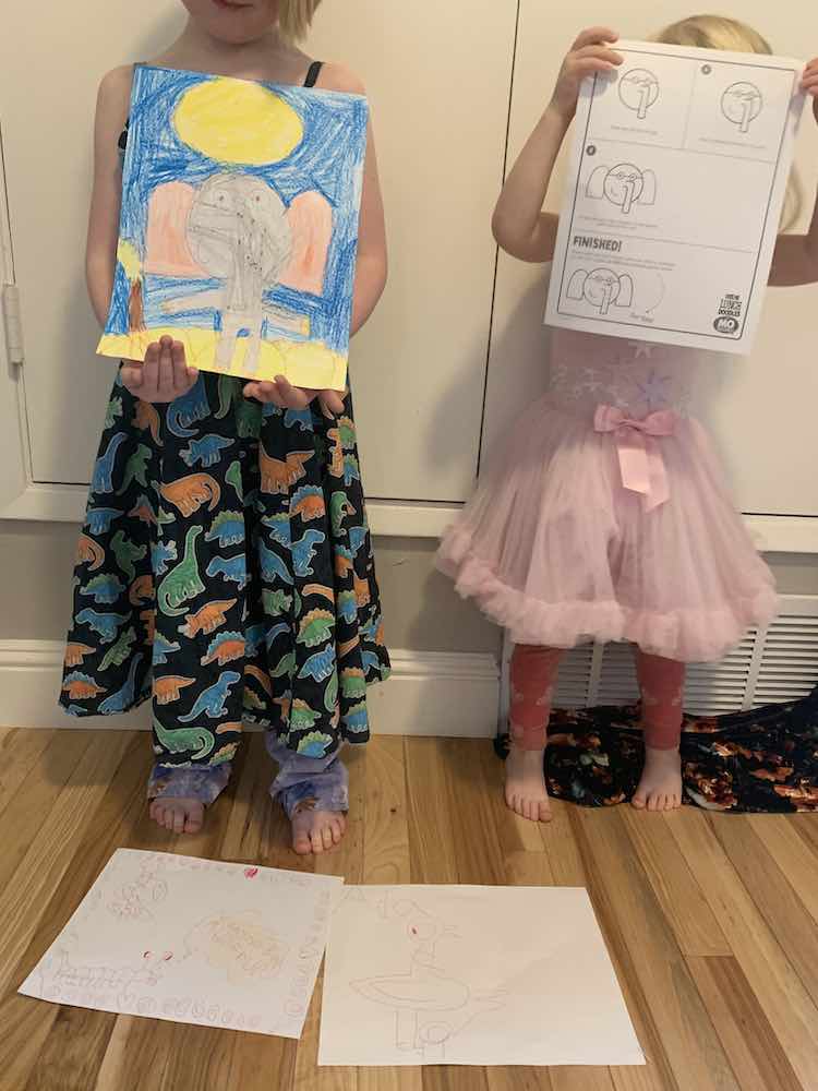 Ada and Zoey standing beside each other. At their feet are two of Ada's doodles on the ground. Ada, on the left, holds her version of Gerald while Zoey, on the right, holds the instruction sheet for Gerald the Elephant.