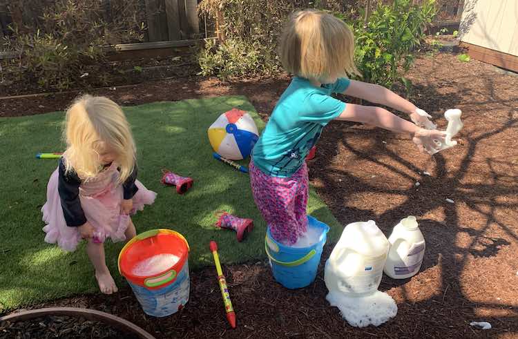 The photo was take outside. To the left Zoey is rolling up her pants about to get in the bucket of soapy water like her sister. To the right Ada is standing in her bucket of soapy water flinging some of the soap bubbles to the right.