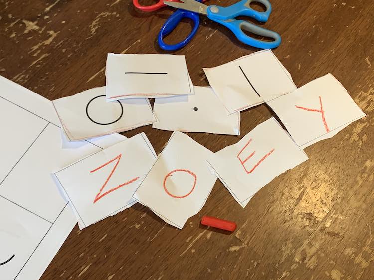 Closeup of the cut out rectangles. The bottom four have the letters Z, O, E, and Y written on them for Zoey's name. Under them is the orange crayon used to write it. Above them are the printed rectangles showing a circle, dot, and a line (one horizontal and one vertical). The scissors and uncut paper are above and to the left. 