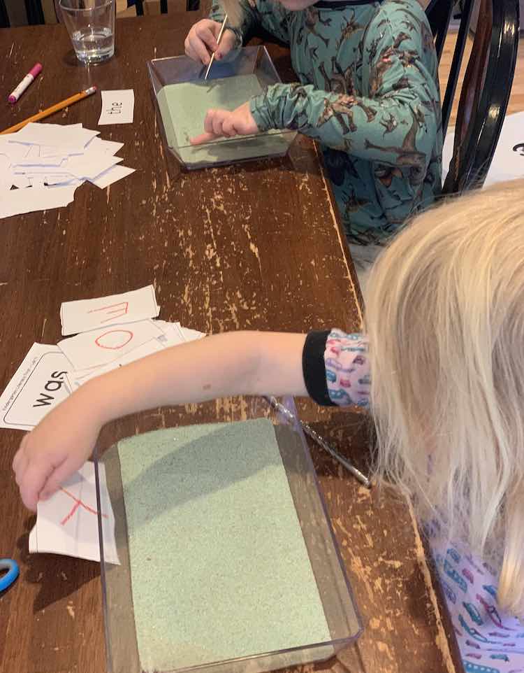 Bottom, forefront, of the image shows Zoey placing the letter 'y' behind the tray so she can sketch it out. In the background, further back, Ada has the 'the' played behind her tray and is drawing it out in her tray.