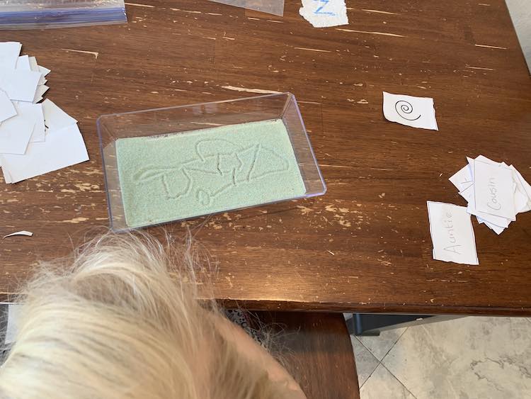 Closeup of Zoey's tray, and top of her head. The tray shows her doodle consisting of lines, a circle, a triangle, and two squares all interconnected. The sight words and shapes are on either side of her tray (hers to the right and Ada's to the left).