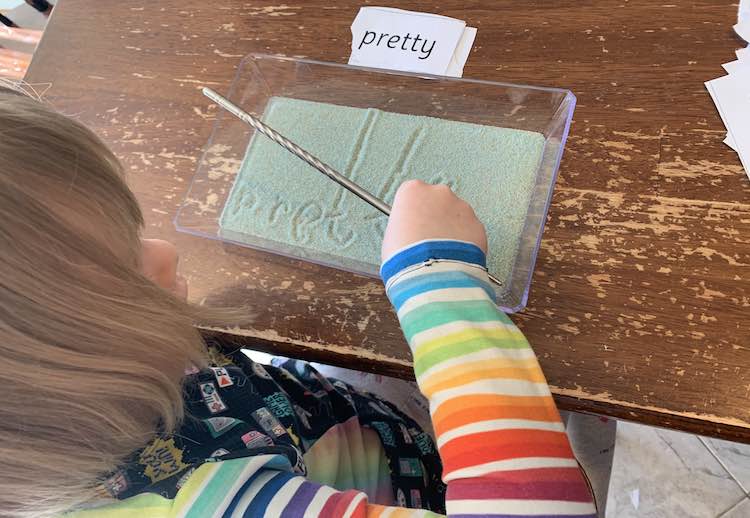 Ada, from above, writing out the word 'pretty'. Her hand obscures the y as she finishes off. The sight word is laid on the other side of the tray with the previously written sight words below it.