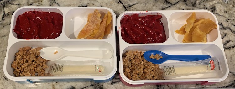 Closeup of both bento boxes showing the contents listed in the caption. Additionally, there's a plastic (Ikea) spoon laid out in the larger hole with the granola and slightly opened cheese string.