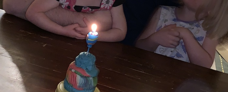 The image shows most of the cake as it sits on a yellow cake platter on the kitchen table. It's pink, blue, and white with a blue balloon candle lit on top. Behind it you can see Ada and Zoey sitting on Matt's lap. 