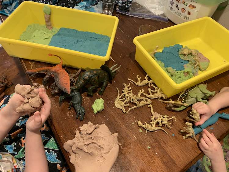 Image shows both girls sitting near each other working on creating a scene in their yellow sensory bins using blue, green, and brown playdough. The dinosaurs are scattered over the table between them and the playdough. 
