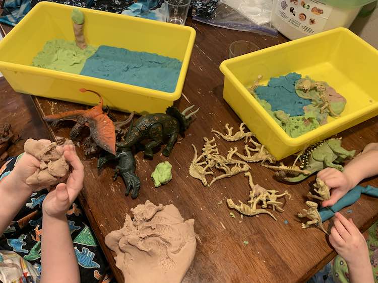 Image shows both girls sitting near each other working on creating a scene in their yellow sensory bins using blue, green, and brown playdough. The dinosaurs are scattered over the table between them and the playdough.