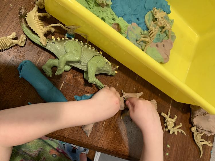 Image shows Zoey shaping some brown playdough in front of her while two skeletons sit inside the bin's scene and several dinosaurs remain scattered around her hands.
