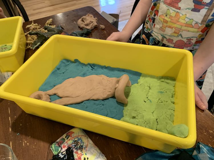 Image shows a closeup of Ada's sensory bin scene while she stands behind it wearing her dinosaur dress. Off to the side you can see more dinosaurs and a small portion of Zoey's bin. 