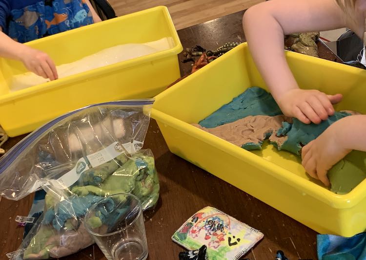 Image shows two bins on the table with a bag of blue, green, and brown playdough between them. The bin furthest away is partially filled with bubbles while Zoey is holding something in it. The bin closest to the camera shows Ada pealing up the layers of playdough from her sensory bin.