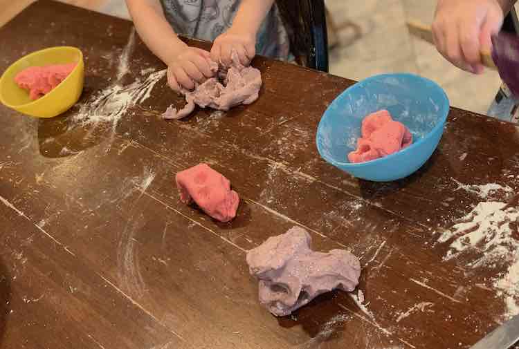 Image is of a floured tabletop. Both girls are sitting down with their glittery playdough in front of them. 