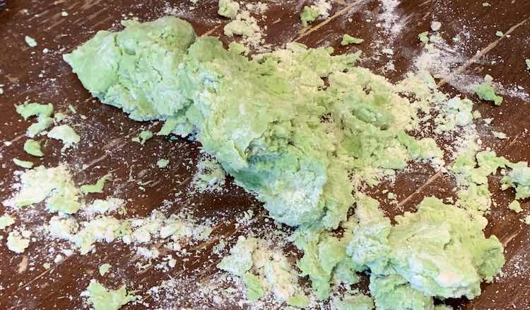 Clumps of green playdough coated, on parts with white. Around the clumps, resting on the table, are lots of white powder from the flour that wasn't able to be mixed in. 
