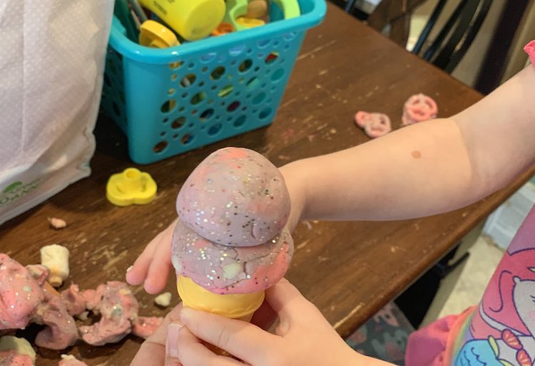 Image shows Zoey holding up a cone filled with two scoops of glittery pink and purple 'ice cream'. The table shows lots of toys in a basket and bag, mounds of the playdough, and some pretzels.