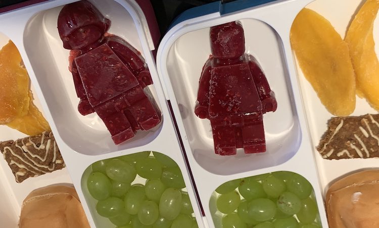 Closeup of the bento boxes side by side. Both LEGO® people (female and male) are shown. The other pockets, slightly cut off, show the dried mango slices, halved Z-bar, bun with cream cheese, and grapes.