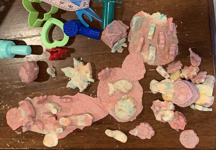 Image is taken from above looking down at the table filled with white mottled cookies placed on flatten pink pieces of Easter egg and bunny imprinted playdough. Off to the side are some of the mentioned tools.