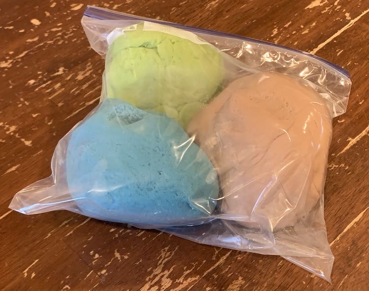 A closed freezer bag lays on the table filled with three colors of playdough. The space between the playdough shows condensation on the inside of the bag since the playdough is still warm.