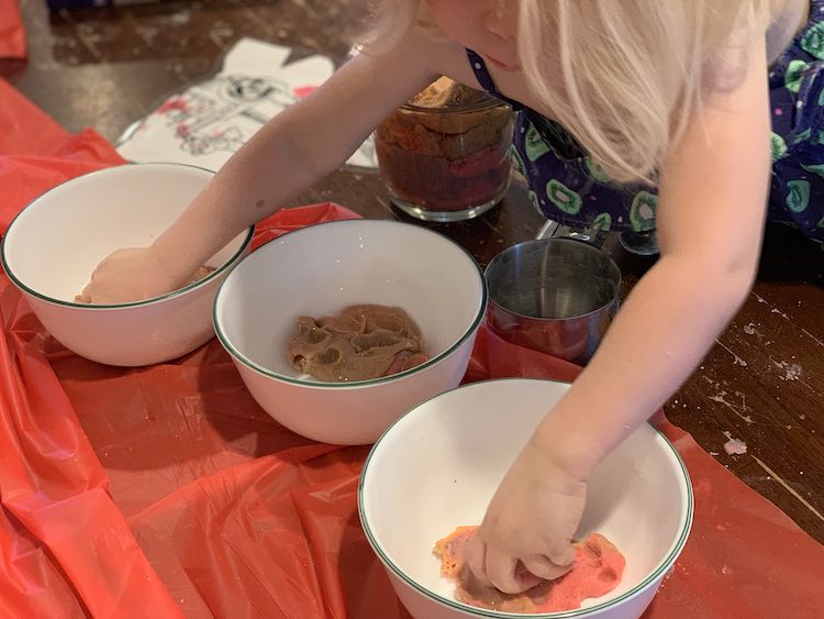 Image shows three white bowls with glittery playdough inside them. Zoey is leaned over the table with one hand in each of the outer two bowls. Under her sits a filled four cup jug with playdough and an empty metal measuring cup. 