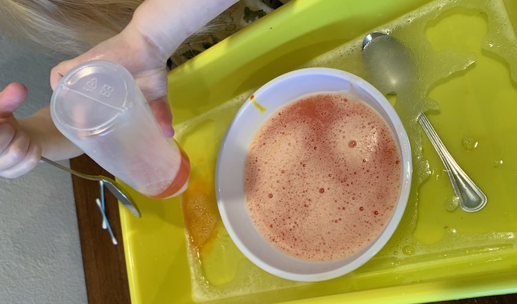 Overview of the yellow bin with vaguely soapy water in it. In the center is a white plastic bowl with soapy orange water in it. Underneath the water you can see a metal spoon, to the right, and orange water spreading, on the left, from where Zoey is squeezing an upside down bottle with orange water in it.