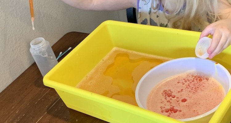 The bowl has been moved to the right side of the bin so you can't see the metal spoon. The bottle has had it's lid removed and now sits lidless to the left. There's a pipette filled with orange water above the bottle and about to be squirted in.