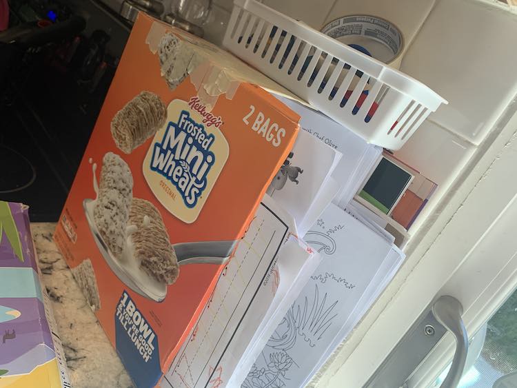 View from the side of the orange boxed paper organizer with the papers already inside it. Above it is a small rectangular plastic basket and in front is the tipped over older cereal box.