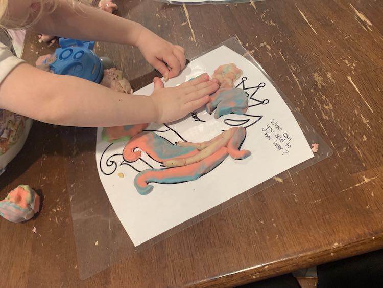 Image shows a laminated unicorn coloring page with the text "What can you add to her hair?". The hair is partially filled in with pastel playdough and Zoey leans over the page pushing some more into it. 