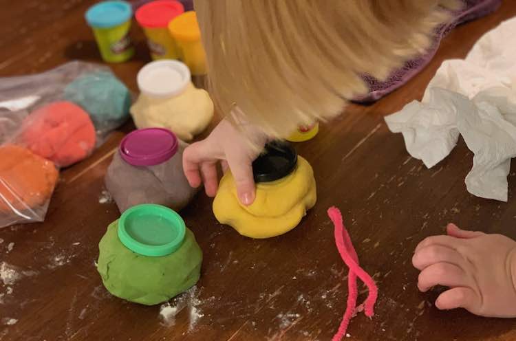 Image shows Ada placing a black lid on the yellow mound of playdough. The green, purple, and white beside it match up. You can see the first colors filled behind and their excess amounts in a clear bag beside it. 