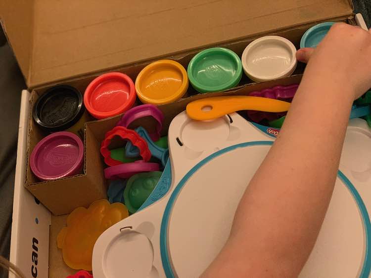 Image shows a cardboard box filled with seven containers of playdough, a white base, and several Play-Doh moulds and cutters. 