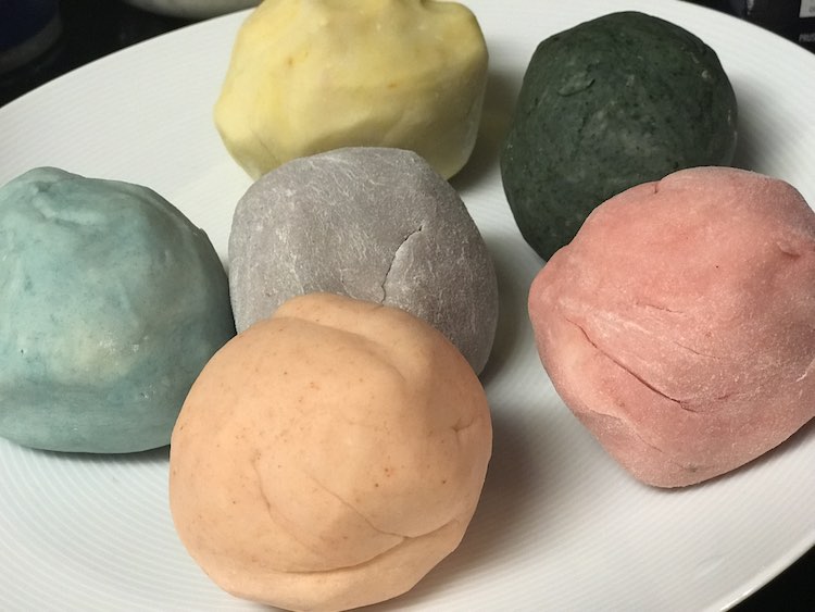 The photo shows a closeup of a white plate. On the plate rests five balls of playdough where one rests in the center and the other five surround the first one.