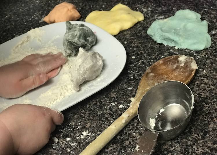 Image shows little hands holding a plate and flattening some pink playdough on a white plate coated in flour. There's two other balls of playdough on the plate with the pink playdough along with three different squished balls behind the plate and a wooden spoon and metal measuring cup beside the plate. 