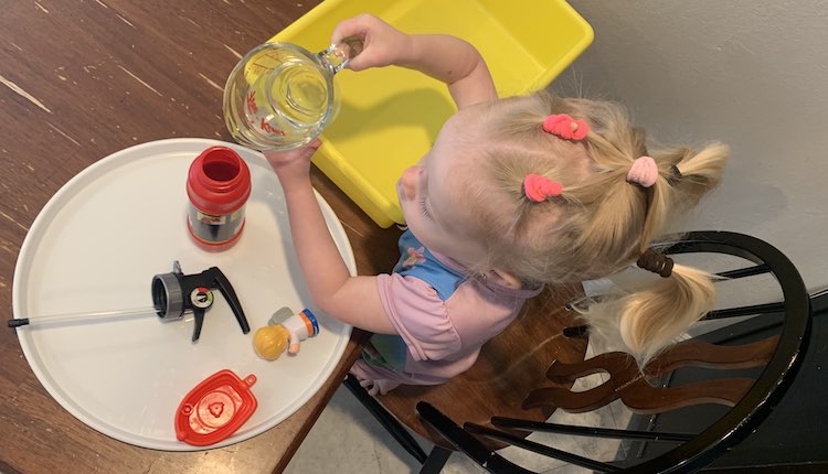 Overhead view looking down at Zoey about to fill the opened fire extinguisher on the white plastic tray. The lid and two plastic toys (Fisher Price boat and doctor) lay beside it.