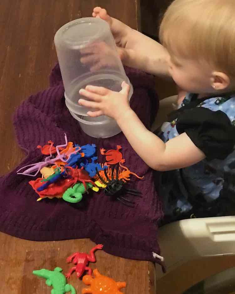 Zoey sits in a booster seat playing with two containers while her right foot rests beside it on the table. Under the containers is a kitchen towel with a pile of plastic animals. The end of the towel closest to use is extremely wet.