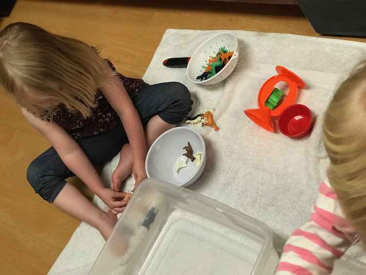 To the left of the photo Ada sits looking down at the plastic characters in her hands. Beside her rests a bowl and handled strainer holding more plastic animals. Almost off photo to the right is Zoey. Between them rests a bin.