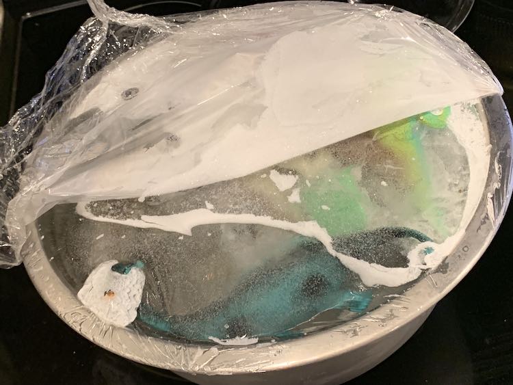 Closeup of the top of the frozen metal bowl with the plastic wrap half pulled off. If you look in the ice you can see a brontosaurus and an green-ish iguana in the depths. 