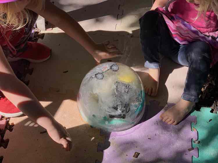 The icy ball is shown placed onto the foam squares outside. Both girls are clustered around it. You can see the odd wheel from the vehicles within.