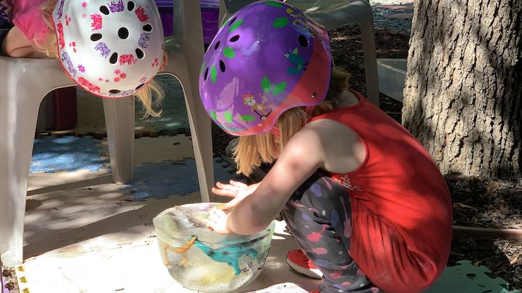 The icy toys are flat side up while Ada rubs the top to try to see what's inside. Zoey is laying on her stomach on the chair looking down at it. From the side you can see the dinosaurs 'running' through the icy side. 