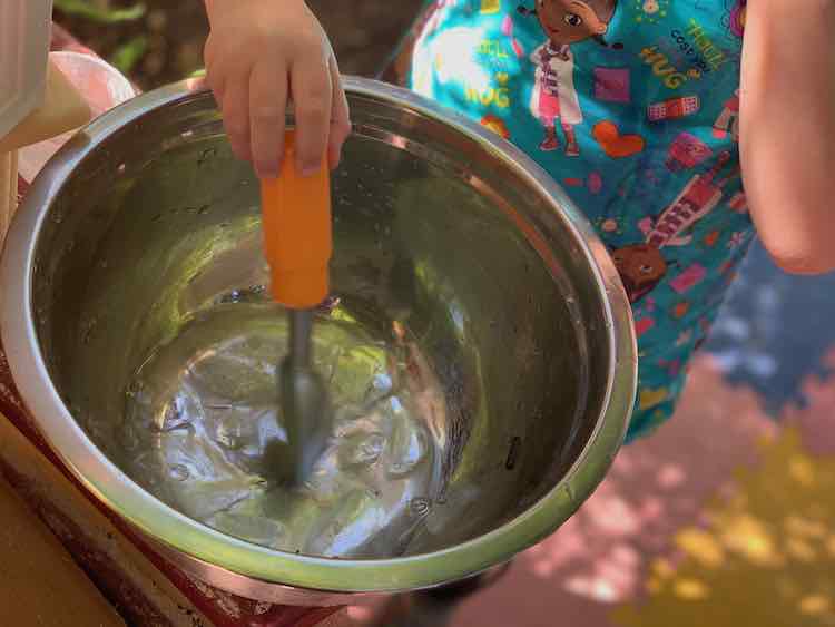 Closeup of the metal bowl with ice and water at the bottom of it. Ada stands behind it and is using a play ice cream scoop to stir the pot. 