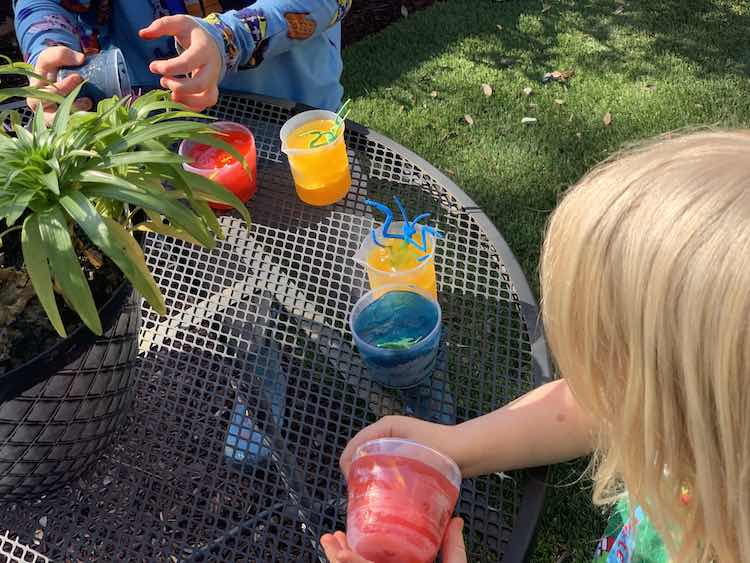 Closeup of part of an outdoor table with the six colored cups lined up. Zoey is closer to the camera holding the red ice cup while Ada is furthest away getting the blue ice out of the cup.