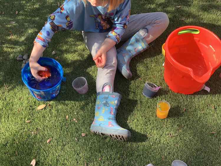 Overview of Ada sitting of fake grass with two buckets of water on either side. She has the filled blue and yellow cup near her leg and the emptied red cup on the other side. She is holding the red ice right above the water in the smaller blue bucket. 