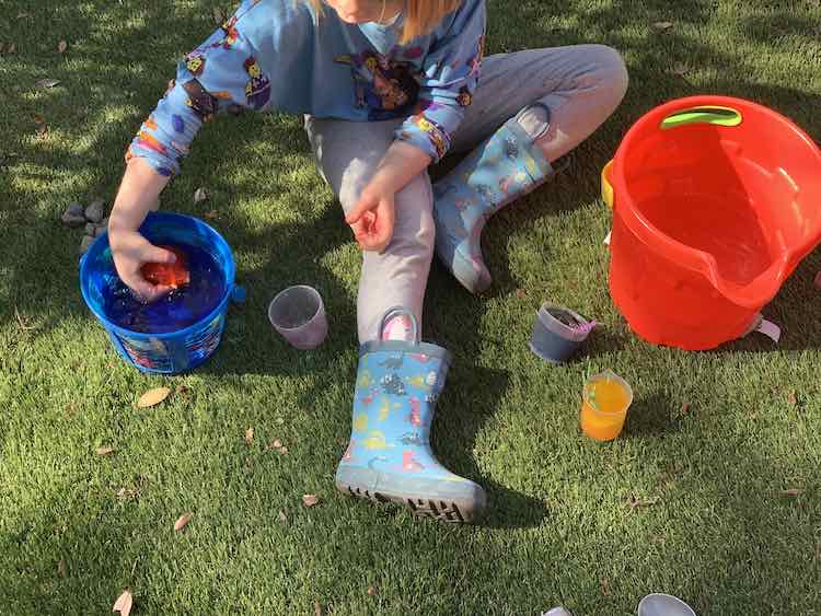 Overview of Ada sitting of fake grass with two buckets of water on either side. She has the filled blue and yellow cup near her leg and the emptied red cup on the other side. She is holding the red ice right above the water in the smaller blue bucket.