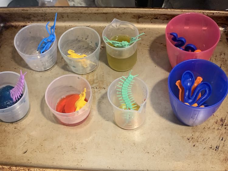 Cookie sheet showing the remnants from before and two new cups with plastic toys. The white cups show the bugs and some of the leftover colored water and un-melted ice.
