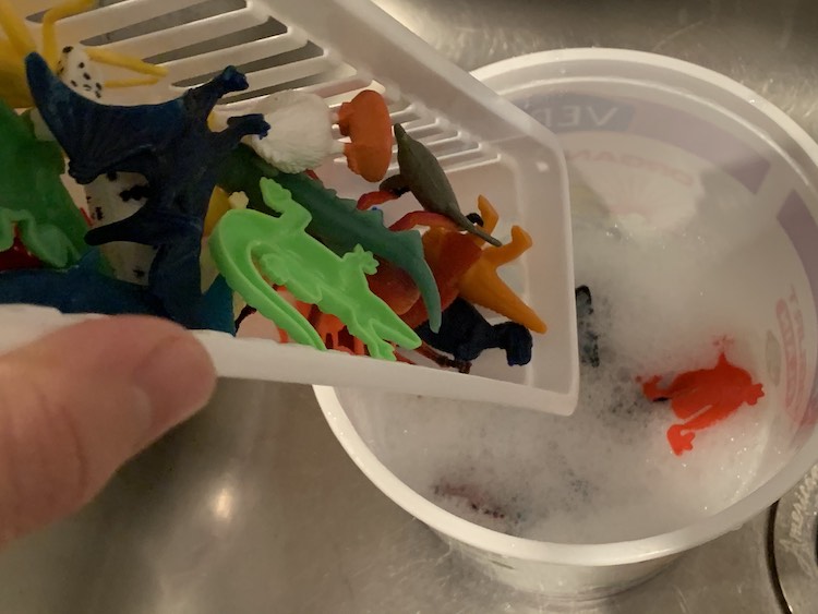 A long plastic holey basket is held tipped over a large yogurt container with soapy water inside. The plastic container holds plastic animals and you can see some of them already in the soapy water below.