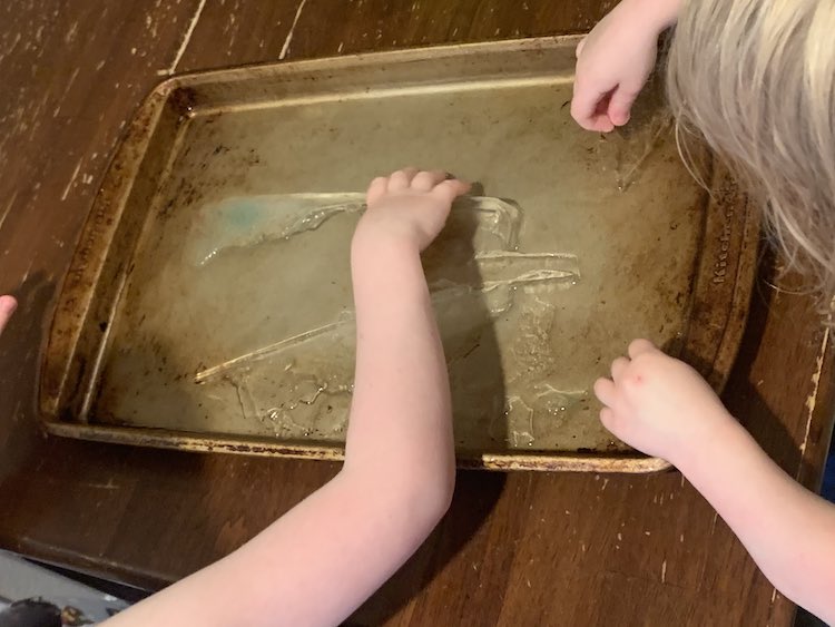 Overview of the cookie sheet on the kitchen table. There's ice frozen to it and the kids are leaned over it picking the ice off to play with.