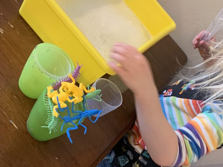 Zoey sitting at the table with a yellow bin full of soapy water in front of her. Beside the bin rests the emptied cups and plastic toys ready to be washed.