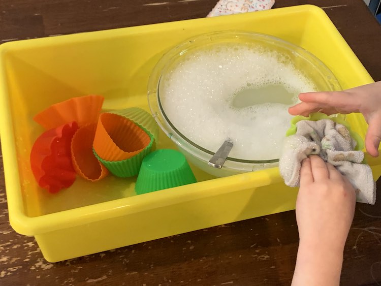 Partially overview of the yellow bin. The right side holds a glass bowl filled with soapy water and a spoon. Over the bowl Zoey's hands are washing a yellow cupcake liner with a small washcloth. To the left, in the yellow bin, a pile of cupcake liners sit waiting to be washed. 