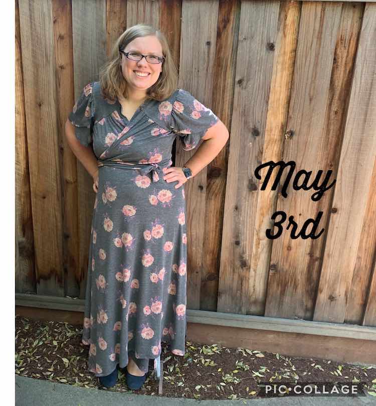 Me standing with my hand on my hips in front of a wooden fence. I'm wearing a grey wrap dress with pink flowers patterned on it. Beside me there's black font saying 'May 3rd'.