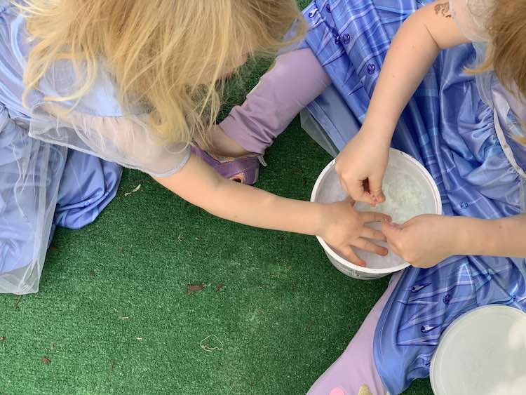 Ada sitting on the fake grass with the container of ice between her legs. She's reaching into the container to play with the frozen bubbles. Zoey crouches beside her and is reaching into the container.