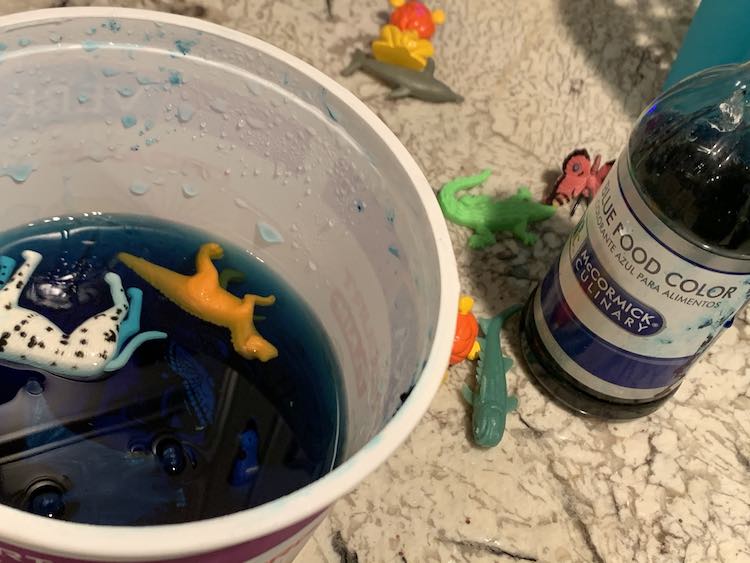 View of the empty yogurt container half filled with blue water and with two animals floating in it. Off to the side is the blue food dye and more plastic animals on the counter. 