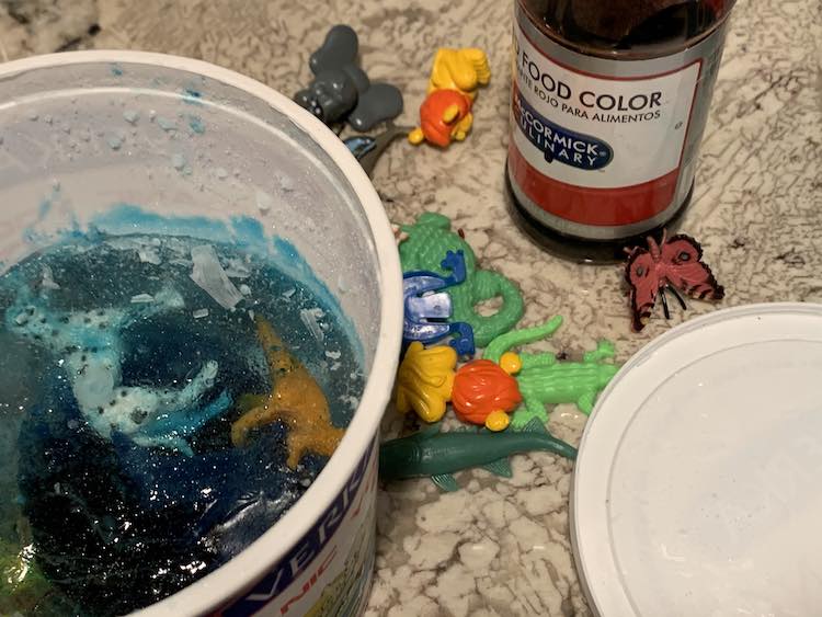 Container of solid blue ice with animals inside it to the right. The left shows the container lid. Behind both are the plastic animals and a bottle of red dye. 