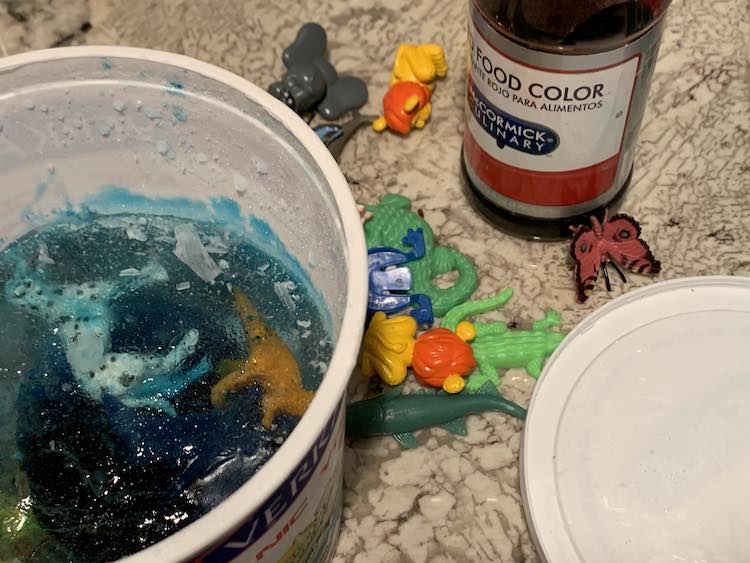 Container of solid blue ice with animals inside it to the right. The left shows the container lid. Behind both are the plastic animals and a bottle of red dye.