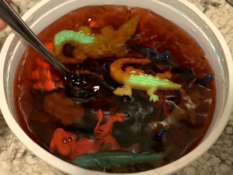 Open container showing toys in red water. There's a spoon handle sticking out and the water is shimmery. 
