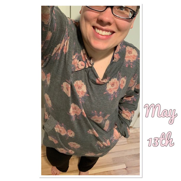 Selfie view with one arm cut off, holding camera, and the other one in my pocket. I'm looking up to the camera wearing a slouchy sweater made out of a thin grey cotton blend with pink flowers on it. To the right of my image are the words, in pink, 'May 13th'.